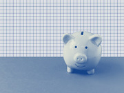 A blue-tinted editorial photo illustration of a smiling ceramic piggy bank against a grid paper background, used as decorative art for a finance-related article.