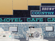 Illustrated or digitally composited scene of Bruno's Country Motel and Cafe exterior with stone wall, signage, and parked vehicles.