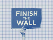 A hand holds a blue campaign sign reading Finish The Wall against a grid-paper background, rendered in a duotone blue editorial illustration style.