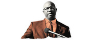 Stylized editorial illustration of a bald man wearing glasses and a brown suit speaking at a microphone, rendered in orange and black tones.