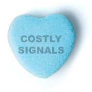 A blue candy conversation heart with the text COSTLY SIGNALS printed on it, used as editorial illustration.