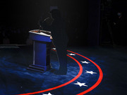 Silhouette of a figure standing at a debate podium on a blue stage with red stripes and white stars, evoking a presidential debate setting.