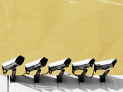 Editorial illustration showing five security surveillance cameras mounted in a row against a yellow background, rendered in black and white.
