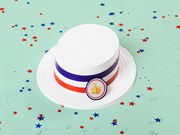 A white boater hat with red, white, and blue stripes and a thumbs-up emoji pin, surrounded by patriotic star confetti on a mint green background.