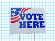 A Vote Here yard sign with American flag motif displayed against a blue grid background, used as editorial illustration.
