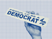 A hand holding a bumper sticker reading Proud To Be A Democrat with the Democratic donkey logo, set against a grid background in blue tones.