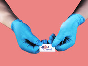 Two hands wearing blue latex gloves hold an I Voted sticker against a coral pink background, illustrating voting during a pandemic.
