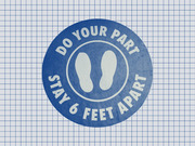 Blue circular social distancing floor sticker with footprint icons reading Do Your Part Stay 6 Feet Apart on a grid paper background.