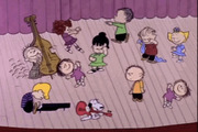 Animated scene from A Charlie Brown Christmas showing Peanuts characters dancing on a stage with Snoopy playing guitar and Schroeder at the piano.