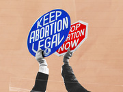 Editorial collage showing two hands holding opposing abortion protest signs: a blue sign reading Keep Abortion Legal and a red stop-sign reading Stop Abortion Now.