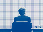 Blue-tinted editorial illustration of a person seen from behind sitting at a desk, overlaid on a grid paper background.